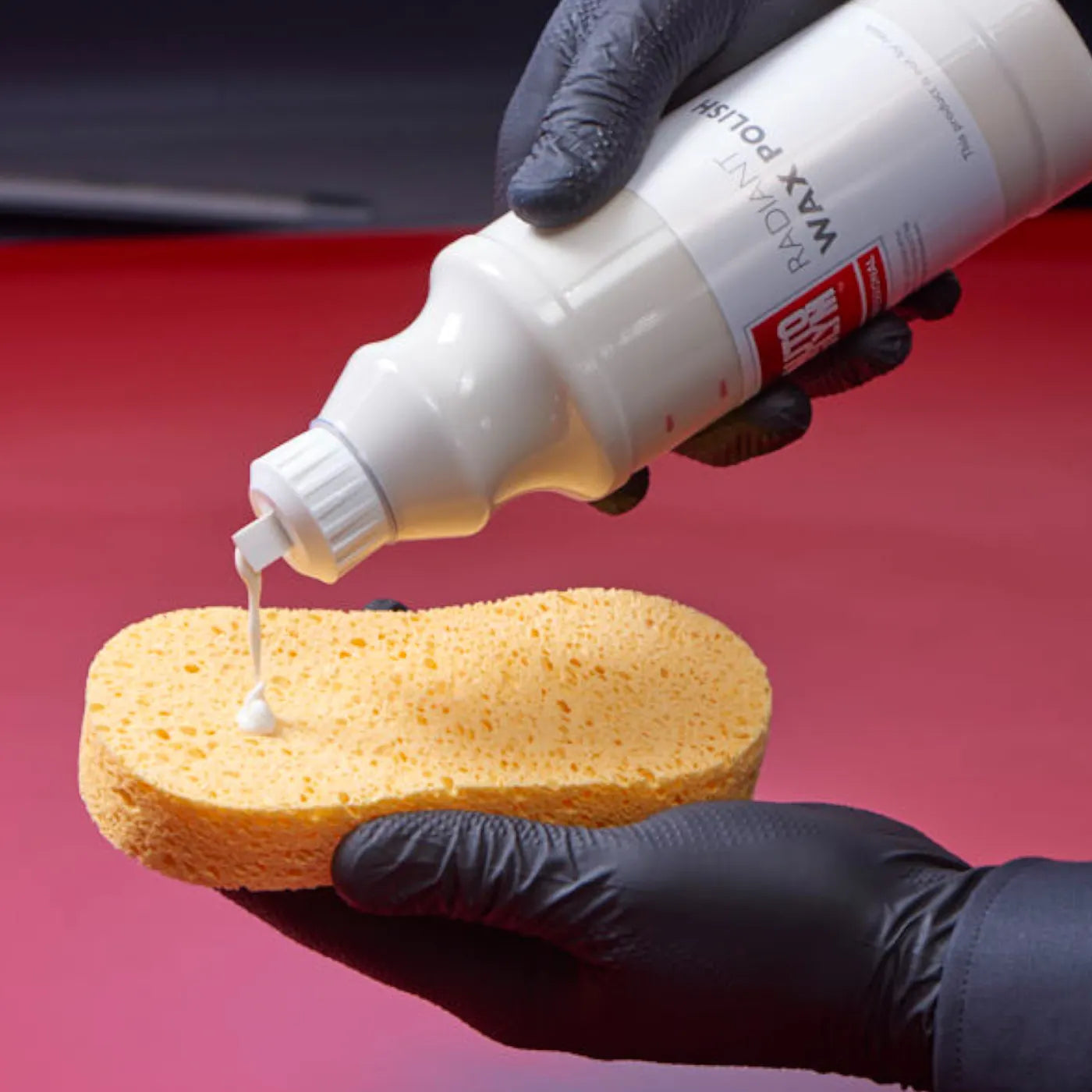 Person applying car wax from a bottle onto a sponge with a red car background