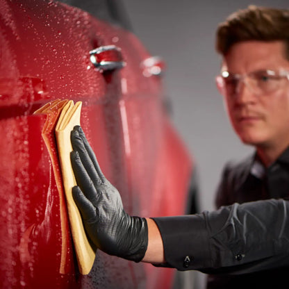 Person cleaning a red car with a yellow towel, wearing black gloves and safety glasses.