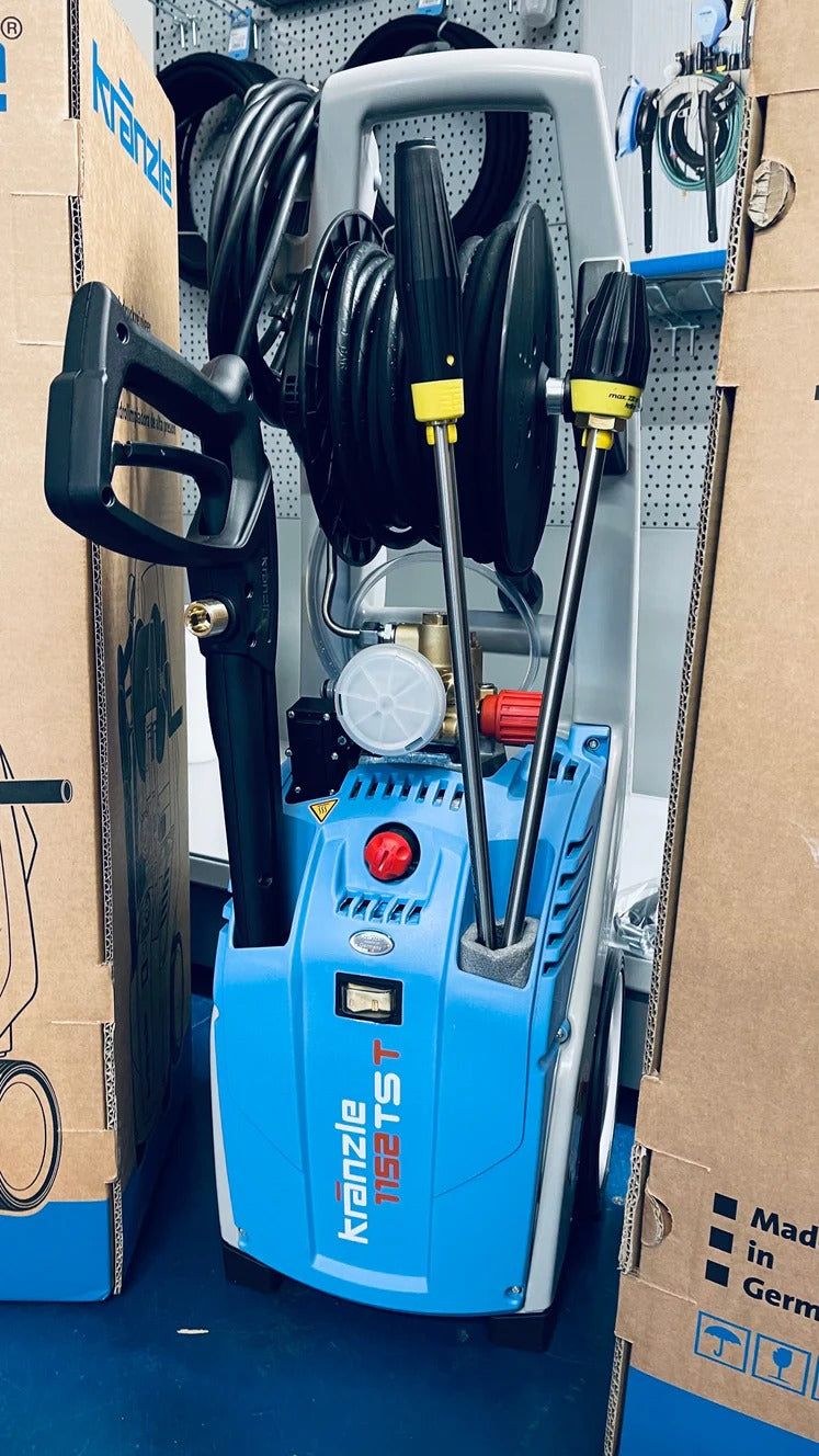 Blue pressure washer with attachments on a blue floor, surrounded by cardboard boxes.